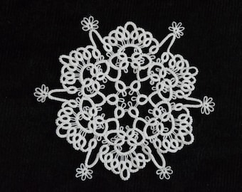 Op de afbeelding: Een witte, ronde doily met een delicaat, kanten ontwerp. De doily heeft een herhalend patroon van lussen en bloemachtige elementen, waardoor een symmetrische, sneeuwvlokachtige uitstraling ontstaat. De ingewikkelde details staan tegen een zwarte achtergrond.