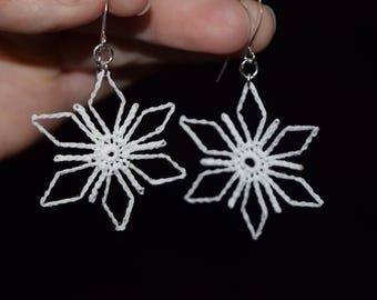 Op de afbeelding: Een paar witte, gehaakte sneeuwvlok oorbellen. Elke oorbel heeft een delicaat, open ontwerp in de vorm van een zespuntige ster. De oorbellen hangen aan zilverkleurige haken tegen een zwarte achtergrond.