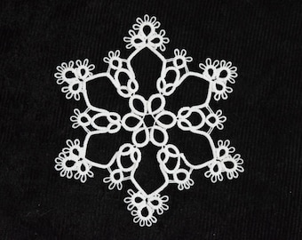 Op de afbeelding: Een delicaat wit sneeuwvlokontwerp tegen een zwarte achtergrond. De symmetrische sneeuwvlok heeft zes punten, elk met gedetailleerde, geluste patronen. Het ontwerp is waarschijnlijk gemaakt van kant of een vergelijkbaar materiaal, wat een gedetailleerd, sierlijk effect creëert.