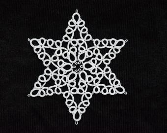 Op de afbeelding: Een witte, stervormige doily met ingewikkelde kanten details, tegen een zwarte achtergrond. Het delicate ontwerp heeft een symmetrisch patroon van geluste en geknoopte draden, wat een feestelijke, handgemaakte uitstraling creëert.