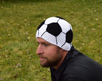 Op de afbeelding: Een handgemaakte gehaakte muts in een voetbalontwerp. De hoed heeft zwart-witte panelen, die het patroon van een voetbal nabootsen. De hoed wordt gedragen door een persoon met een baard, tegen een achtergrond van groen gras.