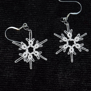 Puede incluir: Un par de pendientes blancos con forma de copo de nieve con ganchos plateados. Los pendientes presentan un diseño calado delicado, que se asemeja a un copo de nieve. Los pendientes están sobre un fondo oscuro.