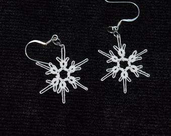Op de afbeelding: Een paar witte sneeuwvlok oorbellen met zilverkleurige haken. De oorbellen hebben een delicaat, open ontwerp dat op een sneeuwvlok lijkt. De oorbellen zijn geplaatst tegen een donkere achtergrond.
