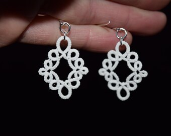 Op de afbeelding: Een paar witte, handgemaakte kanten oorbellen. Elke oorbel heeft een ruitvormig ontwerp met ingewikkelde bloemachtige patronen. De oorbellen hangen aan zilverkleurige oorhaken, tegen een donkere achtergrond.