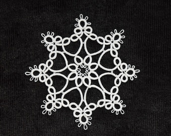 Op de afbeelding: Een witte, handgemaakte sneeuwvlokversiering met een delicaat, kantachtig ontwerp. De sneeuwvlok is gecentreerd op een zwarte achtergrond. De versiering heeft een symmetrische, achtpuntige stervorm, perfect voor winterse feestdagen.