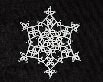 Op de afbeelding: Een wit, handgemaakt sneeuwvlokornament tegen een zwarte achtergrond. De sneeuwvlok is vervaardigd met ingewikkelde kantachtige details, met een symmetrisch ontwerp met bloempatronen. De delicate, open constructie creëert een mooi, luchtig effect, perfect voor winterdecoratie.