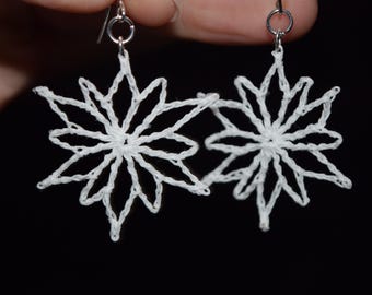 Op de afbeelding: Een paar witte sneeuwvlok oorbellen. Elke oorbel heeft een delicaat, open ontwerp dat lijkt op een ster of bloem. Ze zijn opgehangen aan zilverkleurige oorhaken, waardoor een lichtgewicht en luchtige uitstraling ontstaat. De oorbellen zijn handgemaakt.