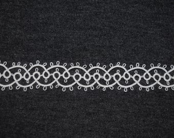 Op de afbeelding: Witte kanten rand met een herhalend bloemmotief tegen een donkergrijze stoffen achtergrond. Het delicate, opengewerkte patroon is geschikt voor diverse knutselprojecten en voegt een vleugje elegantie toe aan kleding of woondecoratie.