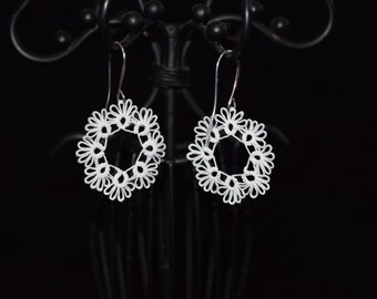 Op de afbeelding: Een paar witte, opengewerkte oorbellen met een bloemmotief. Elke oorbel heeft een cirkelvorm met een centrale opening, omgeven door delicate, bloembladachtige details. De oorbellen hangen aan zilverkleurige haken tegen een donkere achtergrond.