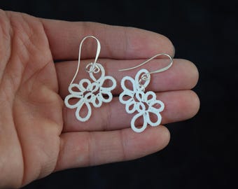 Op de afbeelding: Een paar witte, bloemvormige oorbellen met zilverkleurige haken. De oorbellen worden in een hand gehouden tegen een zwarte achtergrond. De oorbellen hebben een delicate, kantachtige uitstraling.
