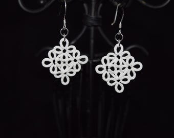 Op de afbeelding: Een paar witte, opengewerkte oorbellen met een bloemmotief. De oorbellen zijn vierkant van vorm met een delicate, kantachtige uitstraling. Ze hangen aan zilverkleurige haken tegen een donkere achtergrond, wat de details van de sieraden benadrukt.