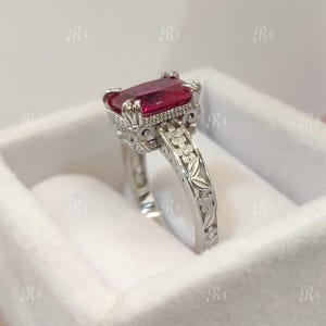Könnte beinhalten: Ein silberner Ring mit einem großen, rechteckigen, roten Edelstein. Der Ring hat ein verziertes Design mit floralen und scrollartigen Details. Der Ring befindet sich in einer weißen Samtbox.