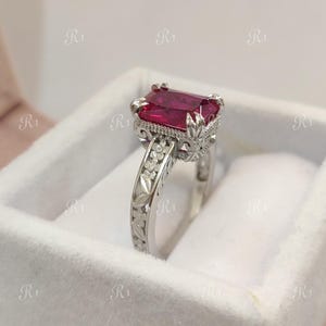 Op de afbeelding: Een zilveren ring met een grote, rechthoekige, dieprode edelsteen. De ring heeft een gedetailleerd, bloemmotief op de band en rond de edelsteen setting. De ring wordt gepresenteerd in een witte fluwelen doos.
