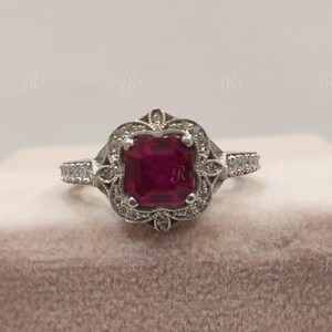 May include: A silver ring with a square, deep red gemstone at the center, surrounded by smaller clear stones. The ring has an ornate, vintage-inspired design with delicate detailing and is set on a pink surface.
