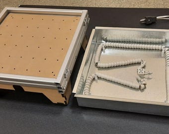 Puede incluir: Una caja metálica rectangular abierta con un elemento calefactor y un marco de madera separado con una superficie perforada. El marco tiene un borde de metal plateado y se sujeta con un clip negro. Un cable de alimentación está conectado.