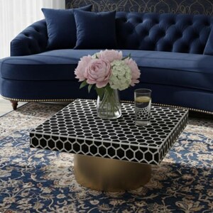 May include: A square coffee table with a black and white honeycomb pattern top and a gold base. A vase of pink and white flowers and a glass of water sit on the table. A blue sofa and patterned rug are in the background.