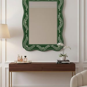 Puede incluir: Un espejo rectangular verde con un borde floral decorativo cuelga sobre una consola marrón. El marco del espejo tiene un borde festoneado y está adornado con patrones blancos en forma de hojas. También se ven una lámpara, frascos de perfume y una pequeña planta.