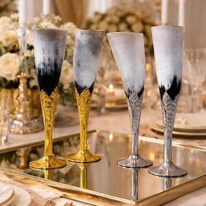 May include: Four champagne flutes with a unique design. The upper portion of each glass features a black and white pattern, while the stems are gold or silver with a leaf-like design. The glasses are arranged on a mirrored tray.