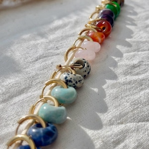 May include: A collection of gold hoop earrings with various gemstone beads, including blue, green, pink, and speckled designs. The earrings are displayed on a wooden stick against a white fabric background.