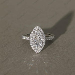 Könnte beinhalten: Ein Diamant-Verlobungsring mit einem zentralen Stein im Marquise-Schliff. Der Ring hat einen Halo aus kleineren Diamanten um den Hauptstein. Das Band ist ebenfalls mit kleinen Diamanten besetzt.