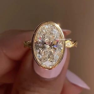 May include: A gold ring featuring a large, oval-cut diamond. The diamond is set in a bezel setting and surrounded by a gold band with intricate detailing. The ring is a piece of jewelry.