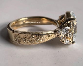 Puede incluir: Un anillo de oro con una piedra central grande y clara de corte rectangular y piedras de acento más pequeñas. La banda presenta grabados florales y el texto "elenajewelryUSA". El anillo está engastado en una banda de oro.