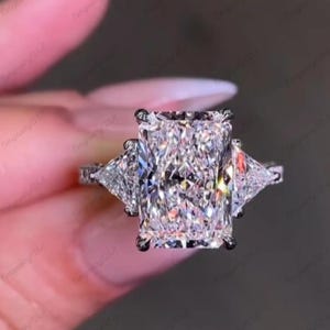 May include: A radiant cut diamond ring with two triangular side stones. The large, rectangular center stone is flanked by smaller, triangular diamonds, all set in a silver band. The ring sparkles with brilliant facets.