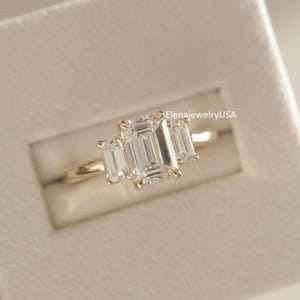 May include: A gold ring with three clear, emerald-cut stones. The center stone is larger than the two side stones. The ring is set in a gold band and is displayed in a light-colored box. The text "ElenajewelryUSA" is visible.