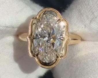 May include: A gold engagement ring with an oval-cut diamond. The diamond is set in a decorative, ornate bezel setting. The ring's band is a simple, polished gold. The ring is displayed on a white surface.