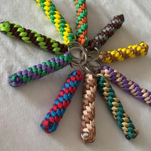 Op de afbeelding: Een verzameling van negen paracord sleutelhangers gerangschikt in een cirkelvormig patroon. Elke sleutelhanger heeft een gevlochten ontwerp in verschillende kleurencombinaties, waaronder geel en groen, oranje en groen, bruin en grijs, paars en groen, rood en blauw, en beige.