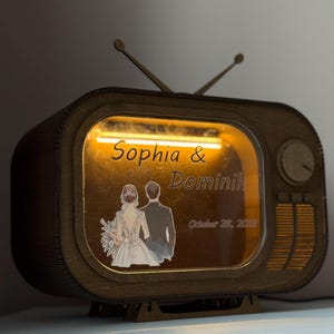 Op de afbeelding: Een donkerbruine houten lamp in de vorm van een vintage televisie met twee antennes. Het scherm toont een warm geel licht, de namen Sophia & Dominik en de datum October 28, 2023, naast een illustratie van een koppel van achteren.