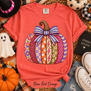 May include: A coral-colored t-shirt featuring a large graphic of a pumpkin decorated with various patterns including flowers, leopard print, ghosts, and lightning bolts, topped with a blue and white striped bow. The shirt has a crew neck.