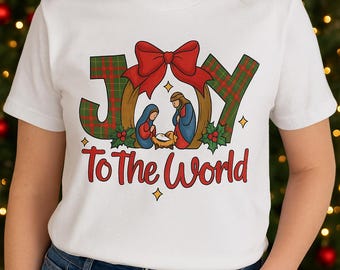 May include: White t-shirt with a Christmas-themed graphic. The design features the word "JOY" in green plaid letters, a red bow, and a nativity scene. The text "To The World" is in red.