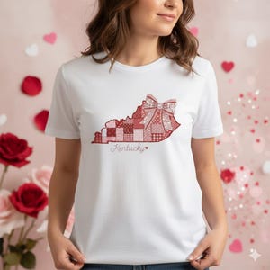 May include: White t-shirt with a red and pink patchwork design in the shape of Kentucky, with a bow and the word "Kentucky" in cursive. The design includes various patterns and a small heart.
