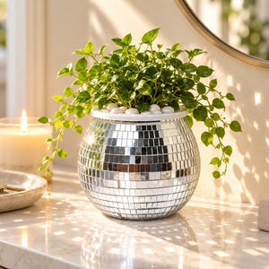 May include: A spherical planter covered in small, reflective silver mirror tiles, containing a lush green plant with small leaves. The top of the planter is filled with smooth white stones, and the surface reflects light in a grid pattern.
