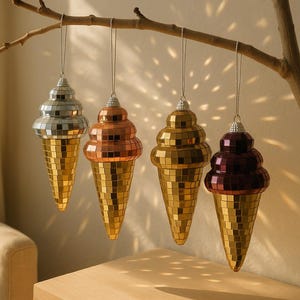 Może przedstawiać: Cztery ozdobne ozdoby w kształcie rożków lodów wiszące na gałęzi. Stożki są złote, srebrne, miedziane i bordowe, z mozaikowym wzorem. Ozdoby zawieszone są na tle ściany w neutralnych odcieniach.