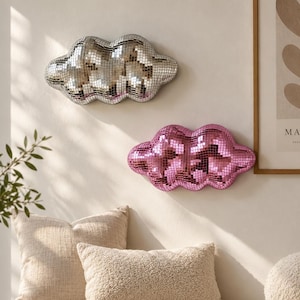 May include: Two cloud-shaped wall decorations covered in small reflective mirror tiles. One cloud is silver and the other is pink. Both items feature a rounded, multi-lobed silhouette and are mounted on a plain wall above a seating area.