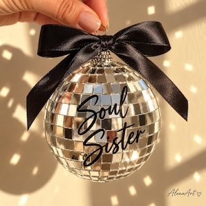 May include: A spherical disco ball ornament covered in small reflective silver mirror tiles. The ornament features the black script text Soul Sister across the center and is topped with a decorative black satin ribbon bow tied at the top.