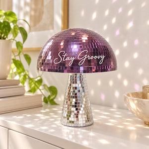 May include: A mushroom-shaped lamp covered in reflective mirror tiles. The dome top features purple mirror tiles, while the base has silver mirror tiles. The text "Stay Groovy" is printed in white script across the front of the purple dome.