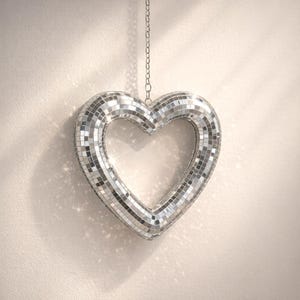Puede incluir: Decoración plateada en forma de corazón, hecha de pequeñas baldosas espejadas. El corazón está suspendido de una cadena y refleja la luz. El objeto decorativo está contra una pared neutra, creando un efecto brillante.