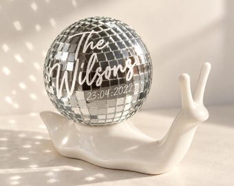 Puede incluir: Una figura de caracol de cerámica blanca con una bola de discoteca plateada reflectante como concha. La bola de discoteca tiene impreso el texto blanco The Wilsons y la fecha 23:04:2022 en una combinación de fuente manuscrita y sans-serif.