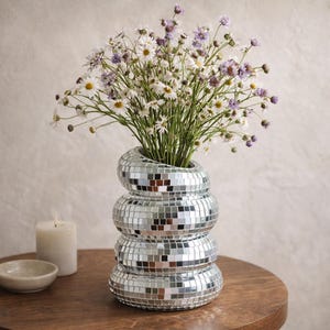 May include: A decorative vase with a mirrored mosaic design, filled with a bouquet of white and purple flowers. The vase is stacked in a unique, rounded shape. A small candle and dish sit on a wooden table.