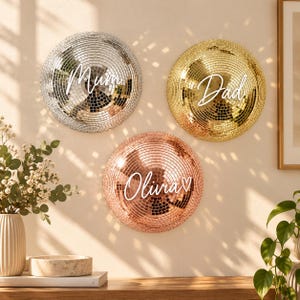 May include: Three circular disco ball wall decorations in silver, gold, and rose gold. The silver ball features the white text Mum, the gold ball features the white text Dad, and the rose gold ball features the white text Olivia with a heart.