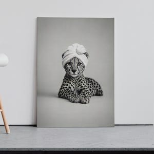 May include: A rectangular wall art print featuring a black and white photograph of a cheetah cub lying down. The cub is wearing a light-colored, textured towel wrapped around its head like a turban. The image is presented in a portrait orientation.