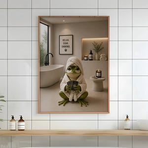 Könnte beinhalten: Ein gerahmter Wandkunstdruck, der einen grünen Frosch in einem weißen Bademantel und einem passenden Handtuch auf dem Kopf zeigt. Der Frosch hält einen kleinen Fächer aus Karten. Der Hintergrund zeigt ein Badezimmer mit einer Badewanne und einem Schild mit der Aufschrift RELAX SOAK UNWIND.