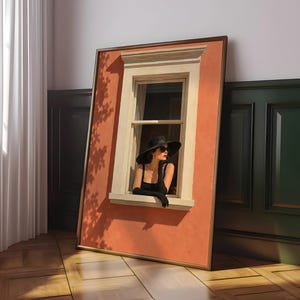 May include: A rectangular framed wall art piece featuring a terracotta-colored wall with a white window frame. A person wearing a black wide-brimmed hat, black sunglasses, and black gloves leans out of the window. The artwork displays tree shadows.