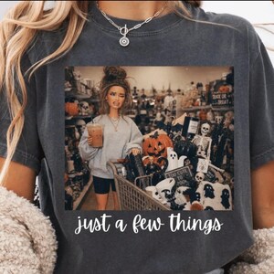 May include: A charcoal gray t-shirt featuring a graphic print of a doll holding an iced coffee in a shopping cart filled with Halloween decorations, including a pumpkin and skeleton, with the white text "just a few things" printed below.