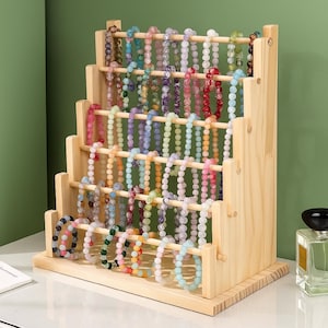 以下が含まれることがあります： カラフルなビーズのブレスレットがたくさん並んだ木製のブレスレットディスプレイスタンド。スタンドは複数の段があり、それぞれにピンク、青、緑、紫など様々な色のブレスレットが並んでいます。スタンドは明るい色の木材で作られています。