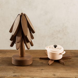 Peut inclure: Un arbre décoratif en bois avec des feuilles ovales et une base ronde. À côté, un petit faitout rose clair avec un bouton noir, posé sur un dessous de plat en bois. Les objets sont sur une surface en bois.