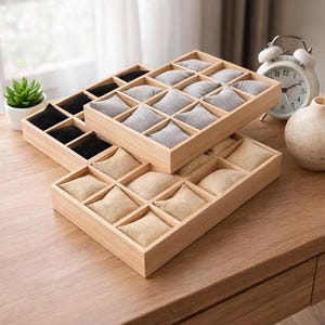 May include: Three wooden jewelry organizer trays with individual compartments. One tray has black cushions, another has gray cushions, and the third has beige cushions. A small succulent plant, a white alarm clock, and a small vase are in the background.
